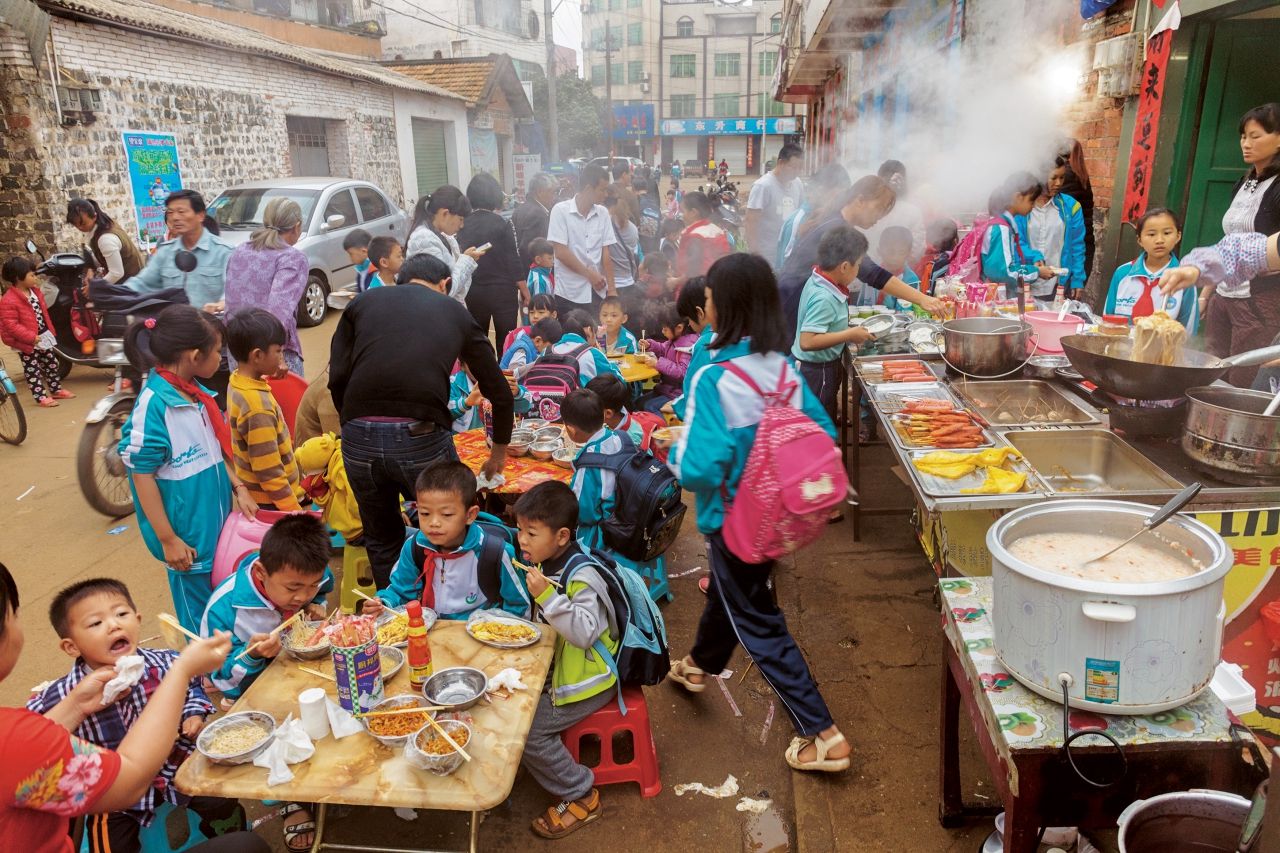 ที่เมืองชวีเจี้ยทางตอนใต้ของมณฑลกวางตุ้ง เด็กๆ กินอาหารเช้าแบบจัดเต็ม ซึ่งมีทั้งบะหมี่ ไข่ และเนื้อสัตว์                                    ที่หน้าโรงเรียน ปัจจุบันหลายครอบครัวทั้งพ่อและแม่ต้องออกไปทำงานนอกบ้าน จึงมีเวลาน้อยลงสำหรับการเตรียมอาหารที่บ้านตามประเพณีดั้งเดิม