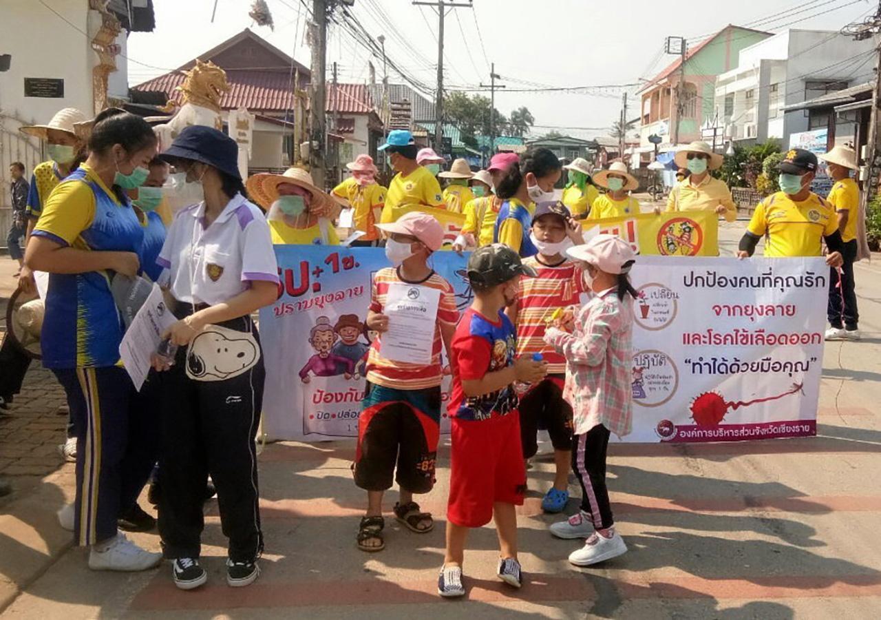 ครูและนักเรียน ถือป้ายออกรณรงค์ให้ชาวบ้านใน อ.เชียงของ จ.เชียงราย ช่วยกันกำจัดยุงลาย ซึ่งเป็นพาหะนำโรคไข้เลือดออก หนึ่งในมาตรการของอำเภอ “คนเชียงของร่วมใจ ปลอดภัยไข้เลือดออก”.
