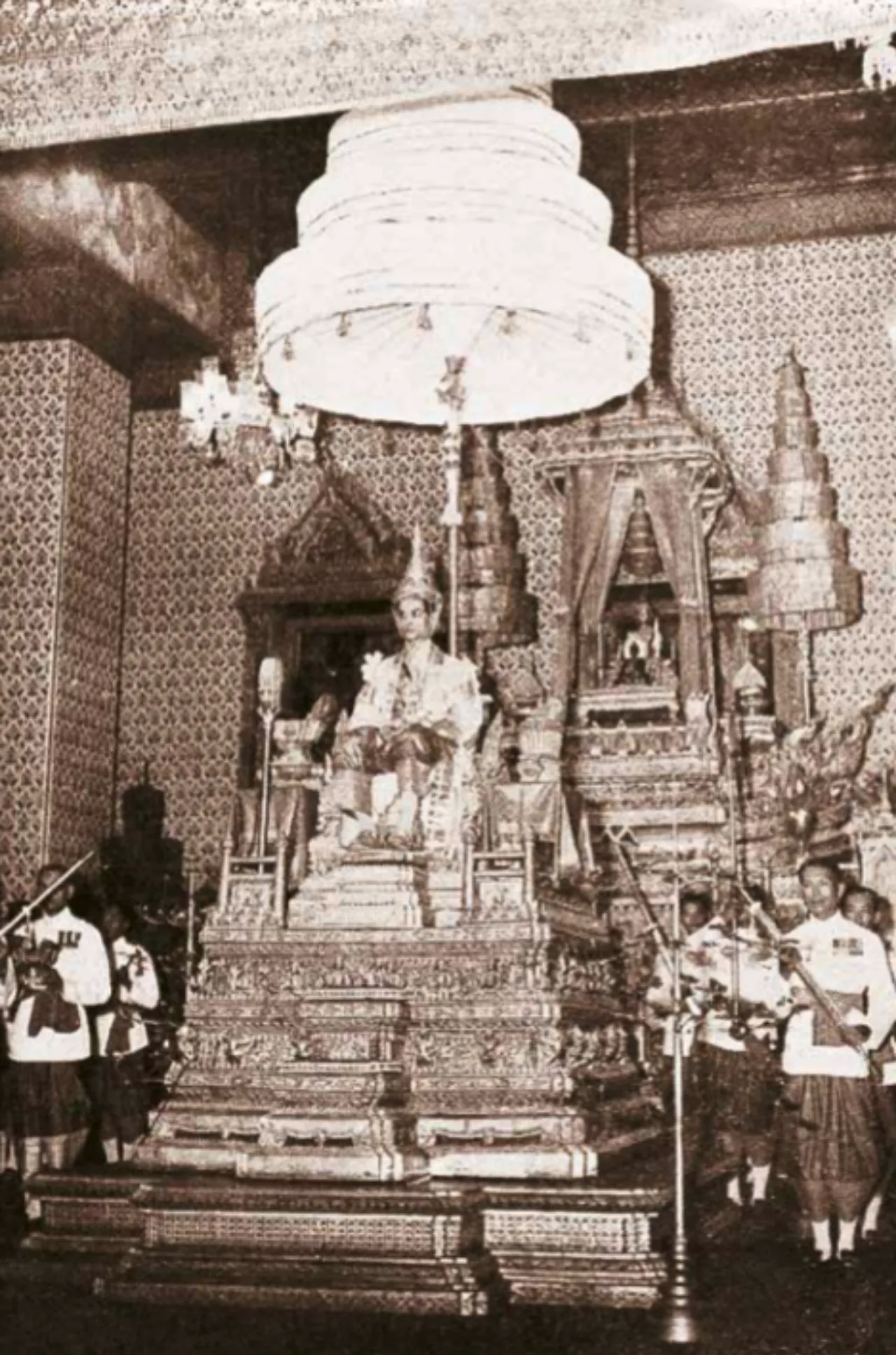พระบาทสมเด็จพระปรมินทรมหาภูมิพลอดุลยเดช บรมนาถบพิตร ทรงเครื่องบรมขัตติยราชภูษิตาภรณ์ ทรงพระมหาพิชัยมงกุฎ ประทับพระที่นั่งพุดตานกาญจนสิงหาสน์ บนพระแท่นราชบัลลังก์ภายใต้นพปฎลมหาเศวตฉัตร แวดล้อมด้วยมหาดเล็กเชิญเครื่องอิสริยราชูปโภคตามตำแหน่ง เสด็จออกมหาสมาคมในการพระราชพิธีบรมราชาภิเษก ณ ท้องพระโรง พระที่นั่งอมรินทรวินิจฉัย
